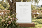 Sage green wedding order of events sign displayed outdoors beside garden flowers, featuring an elegant script heading and minimalist wedding day timeline for a romantic ceremony and reception.