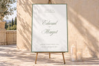 Sage green wedding welcome sign with romantic script names and a fine green border, displayed on an easel beside ivory candles in a sunlit courtyard. Elegant old money wedding entrance signage with soft natural shadows.