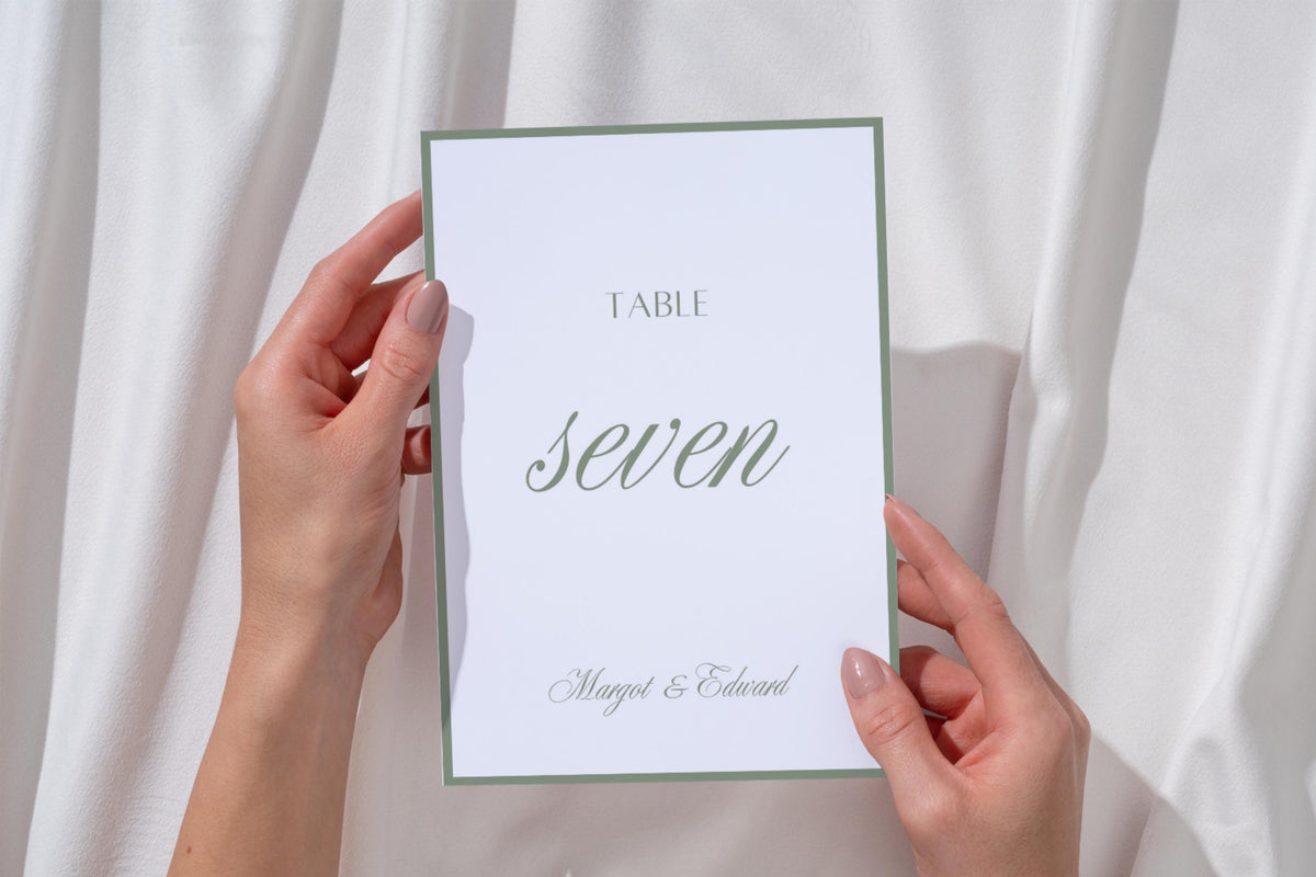 Hands holding a sage green table seven wedding number card against white fabric, showing the elegant script lettering, white background and fine green border of this minimalist wedding reception table sign.