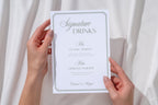Hands holding a sage green Love at First Bite wedding dessert sign with elegant white calligraphy against ivory fabric, showing a close-up of the coordinating reception stationery.