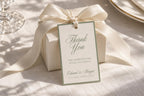 Old money wedding favour tag with sage green border and elegant thank you script, tied to an ivory gift box with satin ribbon, styled on textured linen in natural sunlight.