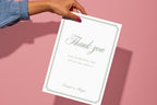 Hand holding a white Thank You for Coming wedding sign with sage green calligraphy and an ornamental border against a pink background, ideal for a wedding favour table.