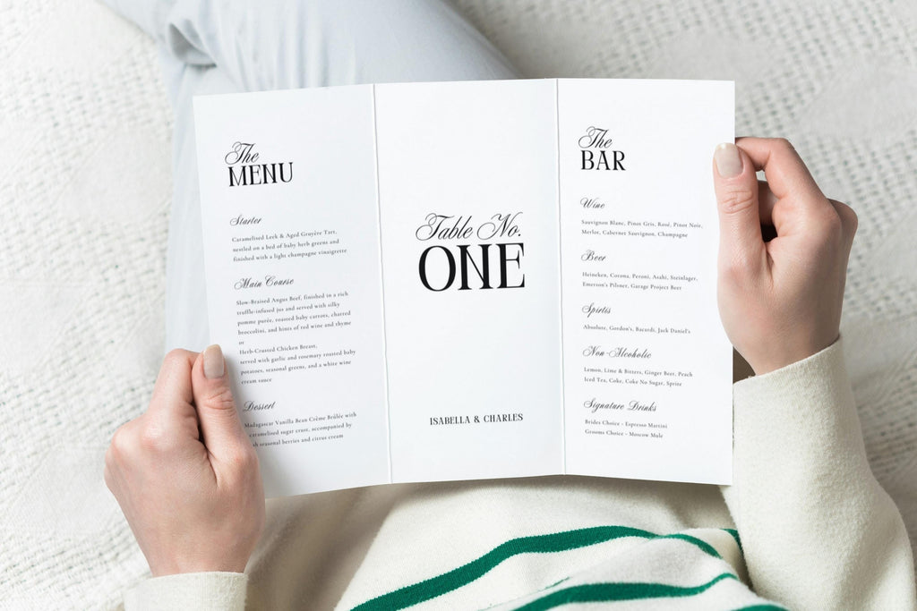 Hands holding an open trifold wedding menu and table number card, editable Canva wedding table decor, refined old money wedding menu and bar signage template