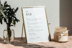 Modern bar menu template for weddings displayed in gold frame with greenery, includes editable drink list, wine, and cocktail sections