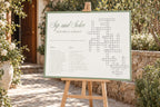 Editable sage green wedding crossword game sign on a wooden easel beside white flowers and greenery at an outdoor garden wedding.