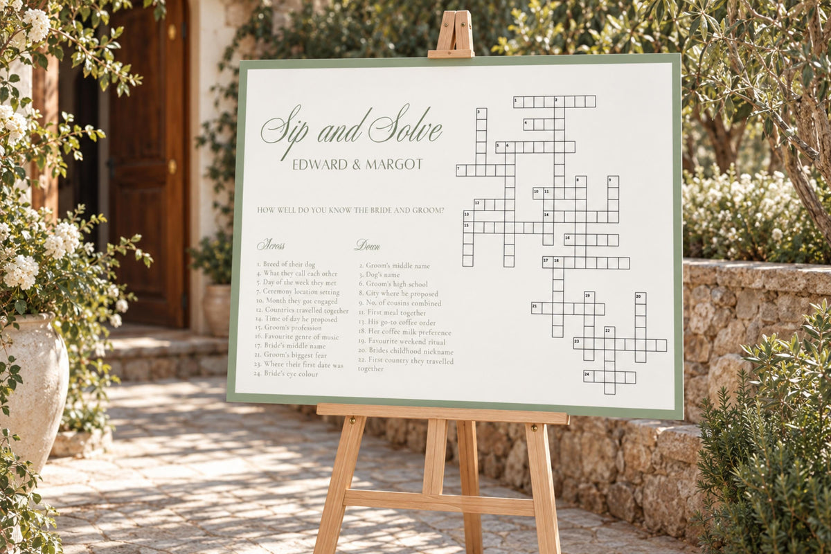 Editable sage green wedding crossword game sign on a wooden easel beside white flowers and greenery at an outdoor garden wedding.