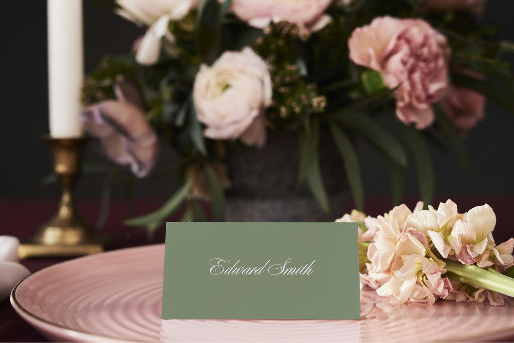Folded sage green wedding name card with elegant white script on a pink dinner plate, surrounded by pastel flowers and candlelight for a romantic old money wedding reception.