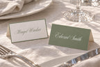 Two coordinating sage green wedding place card designs, featuring an ivory card with a green border and a solid green card with white script, displayed together on a linen reception table.