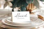 Minimalist folded wedding place card with script and serif font on modern neutral tablescape, editable Canva template