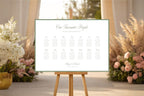Sage green bordered wedding seating chart template displayed on an easel beside pastel flowers at an elegant outdoor wedding venue.