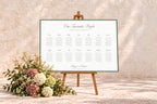 Landscape wedding seating chart with a sage green border displayed on a wooden easel against a warm stone wall, styled with soft neutral wedding florals.