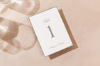 Printable sage green wedding table number card lying on a neutral fabric background with soft sunlight and shadows, showcasing a fine green border and timeless typography for minimalist wedding table décor.