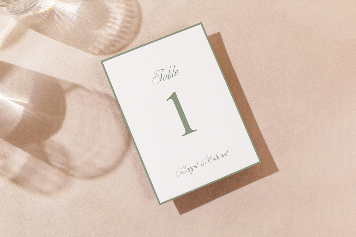 Printable sage green wedding table number card lying on a neutral fabric background with soft sunlight and shadows, showcasing a fine green border and timeless typography for minimalist wedding table décor.