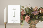 Elegant sage green wedding table number in a delicate metal holder beside a white taper candle and pastel floral centrepiece, featuring classic typography for a romantic garden wedding reception.