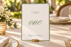 Elegant sage green table one sign in a gold holder on an ivory wedding reception table, featuring romantic calligraphy and a minimalist border for classic garden wedding table decor.