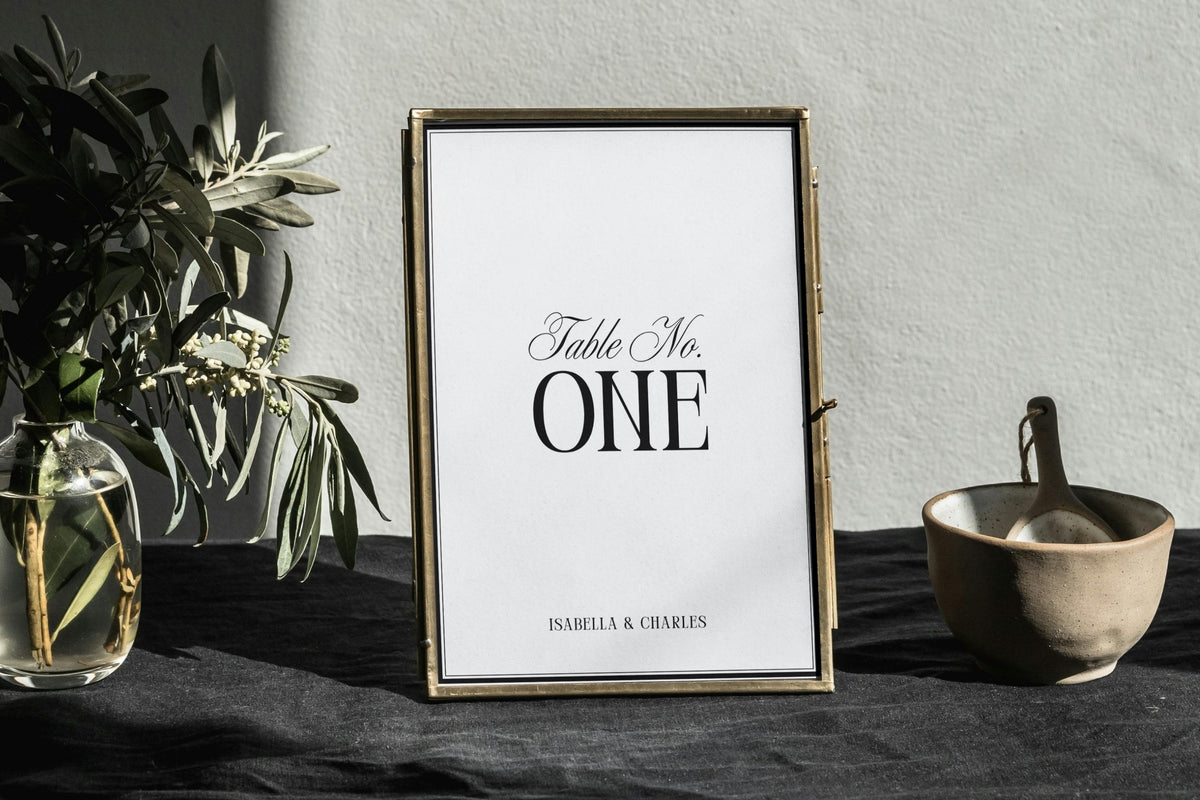 Minimalist wedding table number template displayed in a gold frame on styled table, editable black and white table number design for modern wedding décor, printable Canva wedding signage.