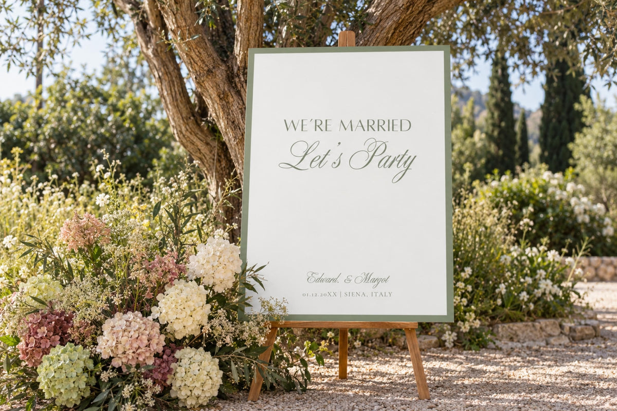 Elegant sage green wedding welcome sign reading “We’re Married, Let’s Party,” displayed on a wooden easel among white flowers and greenery at an outdoor garden wedding.