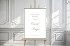 Elegant rehearsal dinner welcome sign on a wooden easel in a light-filled room with decorative wall panelling. White and sage green wedding eve signage with personalised script names and timeless old money styling.