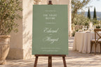 Sage green rehearsal dinner welcome sign with ivory lettering and romantic calligraphy on a wooden easel at an outdoor wedding venue. Elegant “The Night Before” sign for a wedding eve celebration.