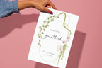Floral wedding guestbook sign held against a pink background, featuring an illustrated green ribbon border and script lettering for a romantic wedding guestbook table.