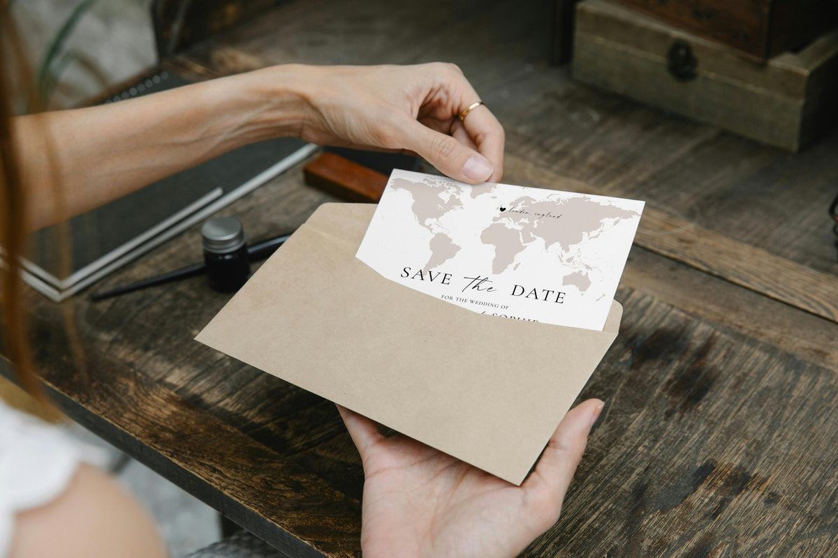Modern travel-themed save the date card being slipped into kraft envelope, minimalist world map design editable with Canva.